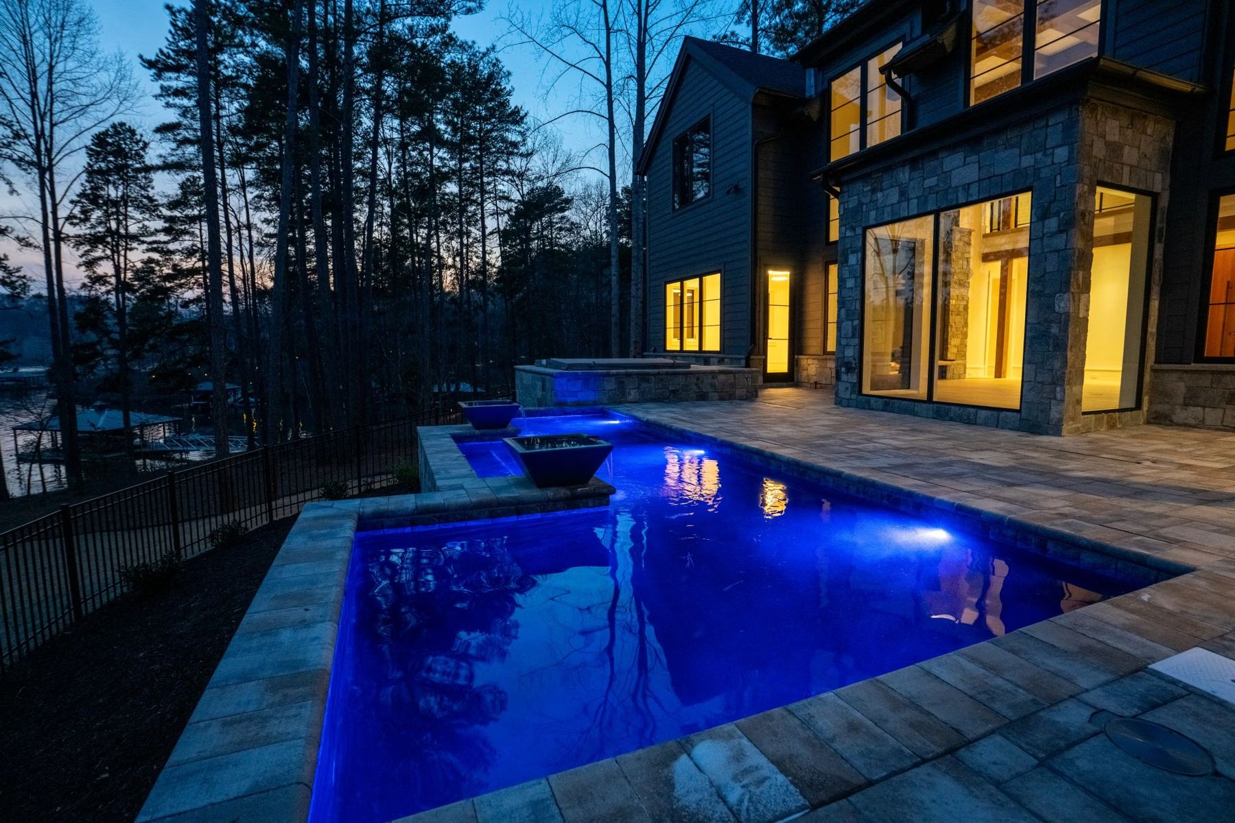 A pool and fire bowls at dusk beside a lake house